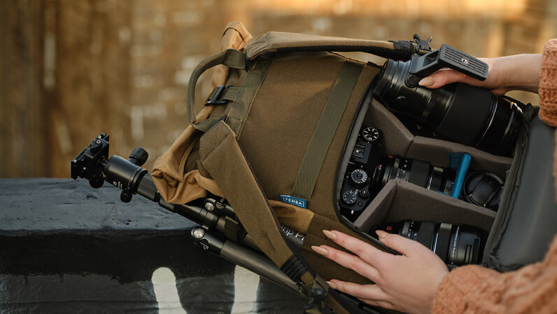View of Tenba Fulton version 2 backpack open with multiple accessories fit inside. Accessories include a mirrorless camera with attached lens and two additional lens with a tripod in the exterior pocket on the side of the backpack.